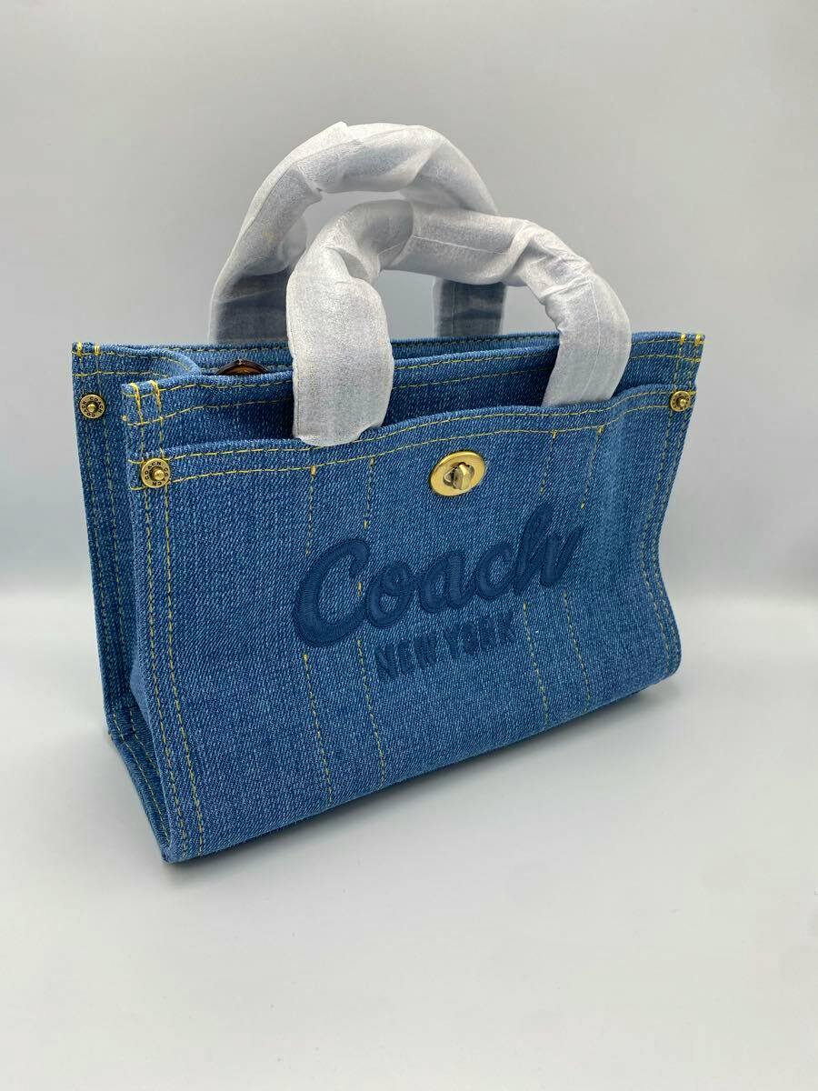 Three-quarter photo of the blue denim Coach tote bag showing the Coach logo on the front, gold turn-lock closure, and handles wrapped in protective paper.