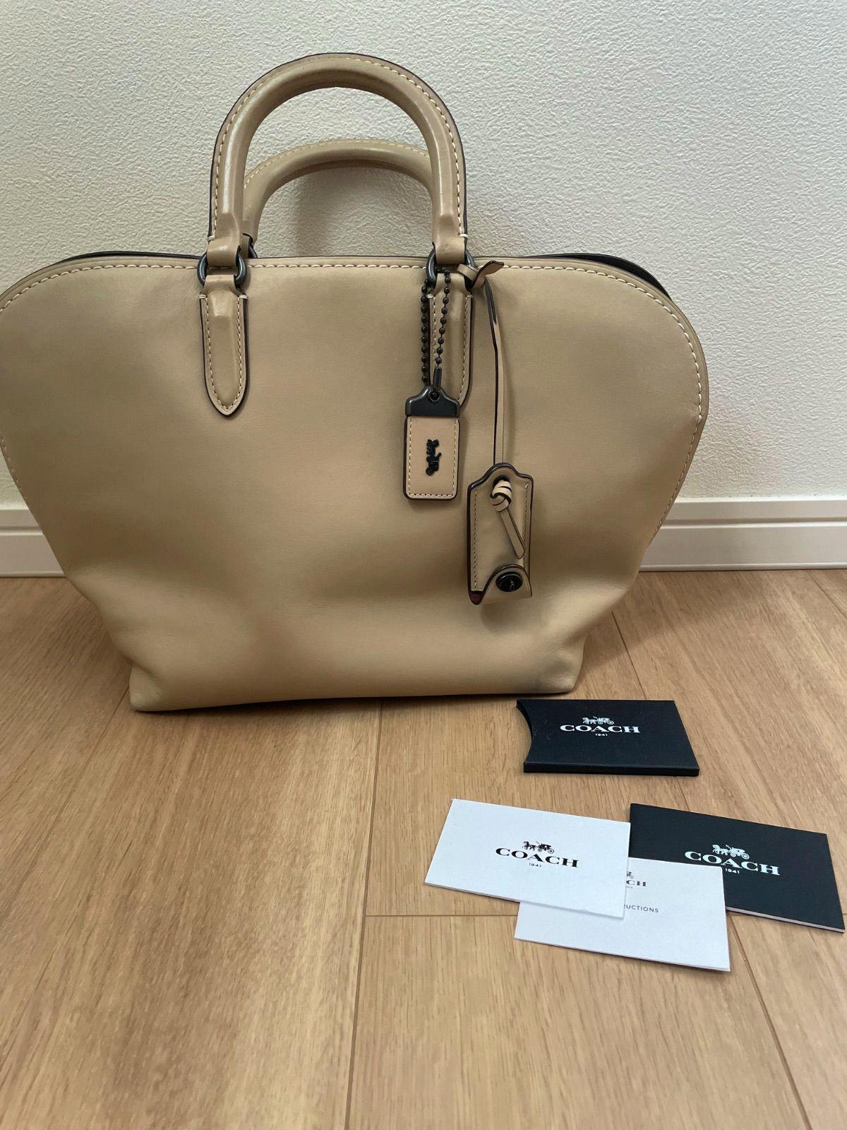 Beige COACH Dakota 2-way leather satchel handbag shown upright with attached leather tags and Coach booklets on a wooden floor.