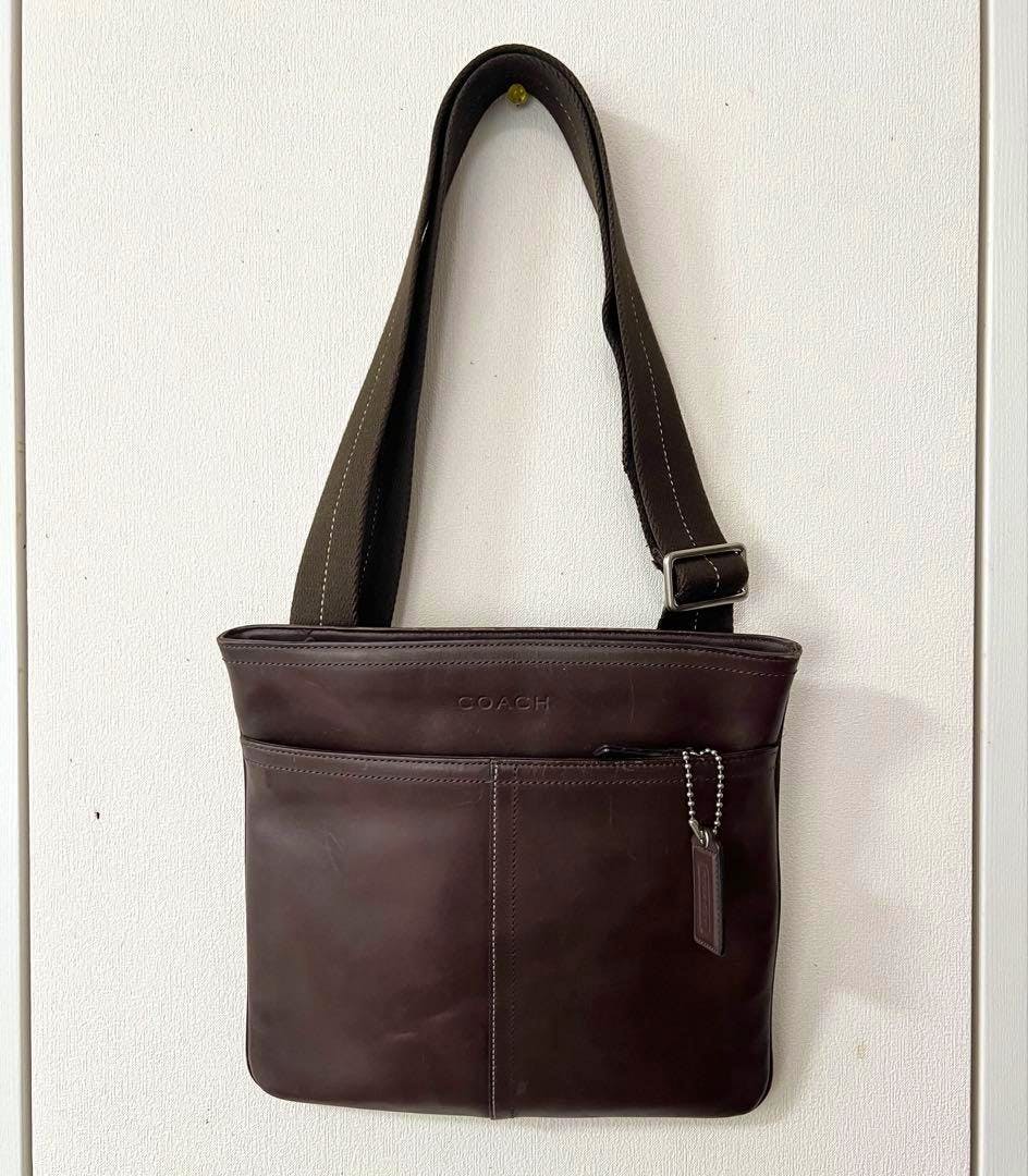 Dark brown Coach leather shoulder/body bag hanging on a wall, front view showing exterior pocket and leather tag.