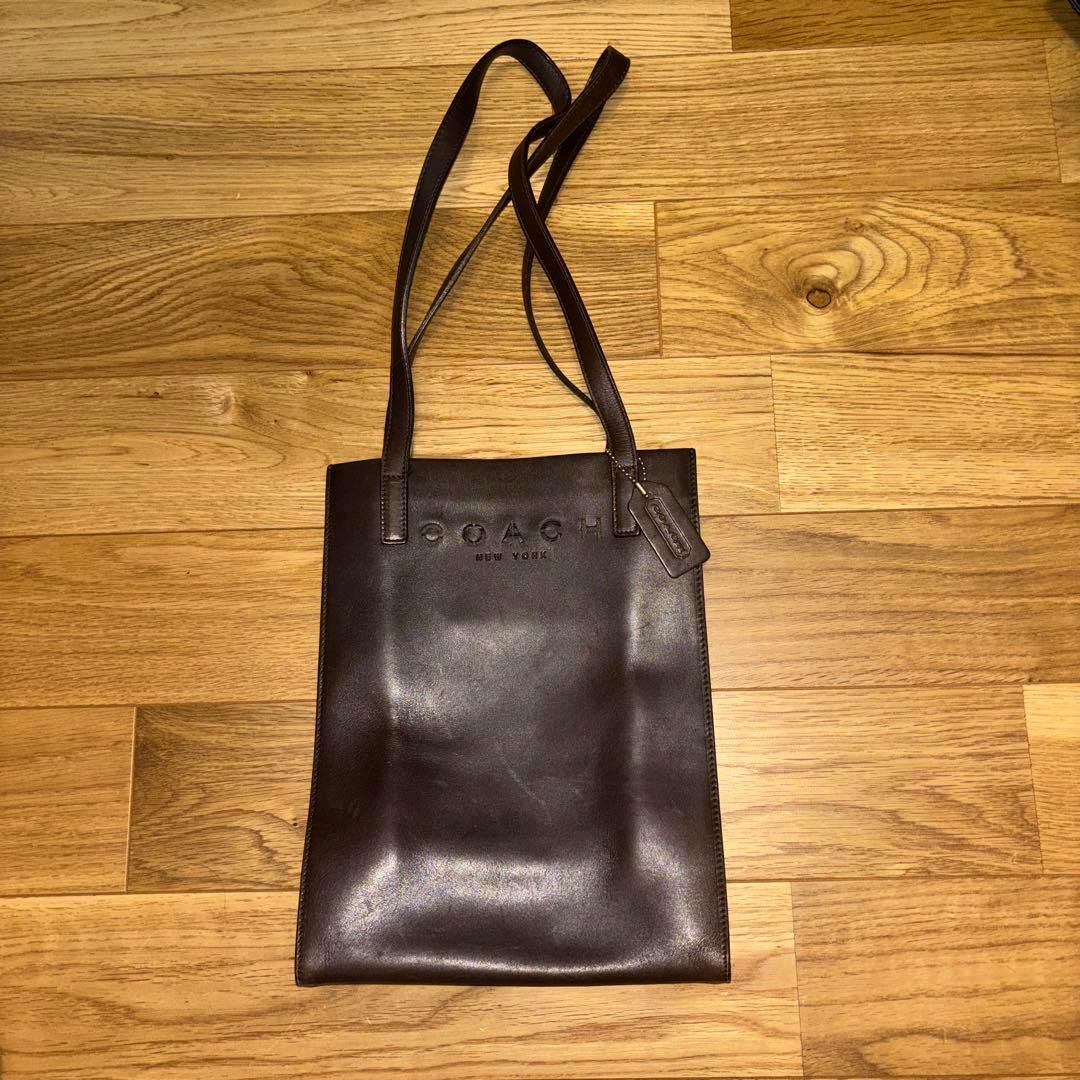 Dark brown cow leather Coach shoulder bag with long straps and an embossed "COACH NEW YORK" logo lying on a wooden floor.