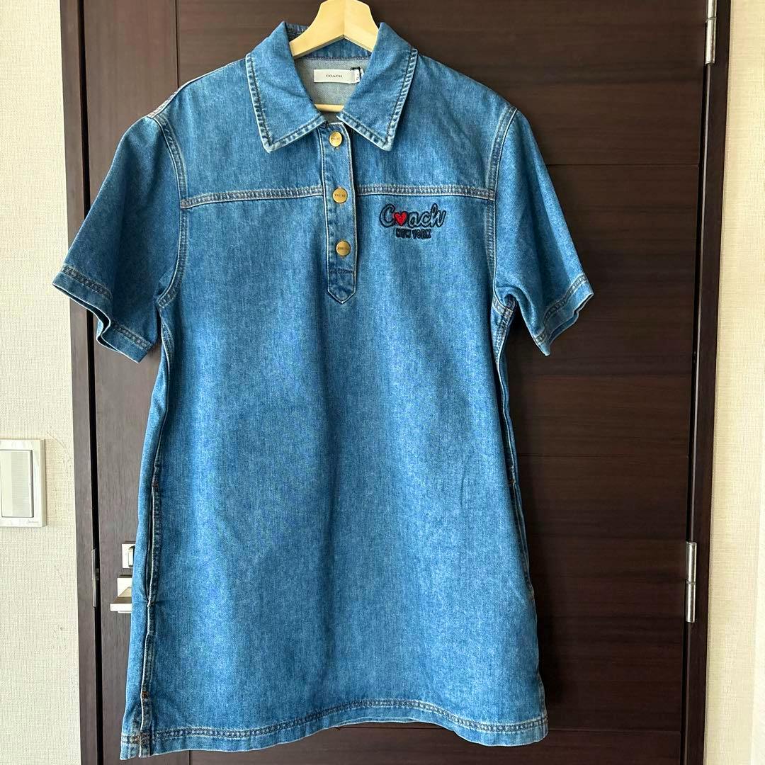 Blue denim Coach shirt dress hanging on a wooden hanger against a dark door.