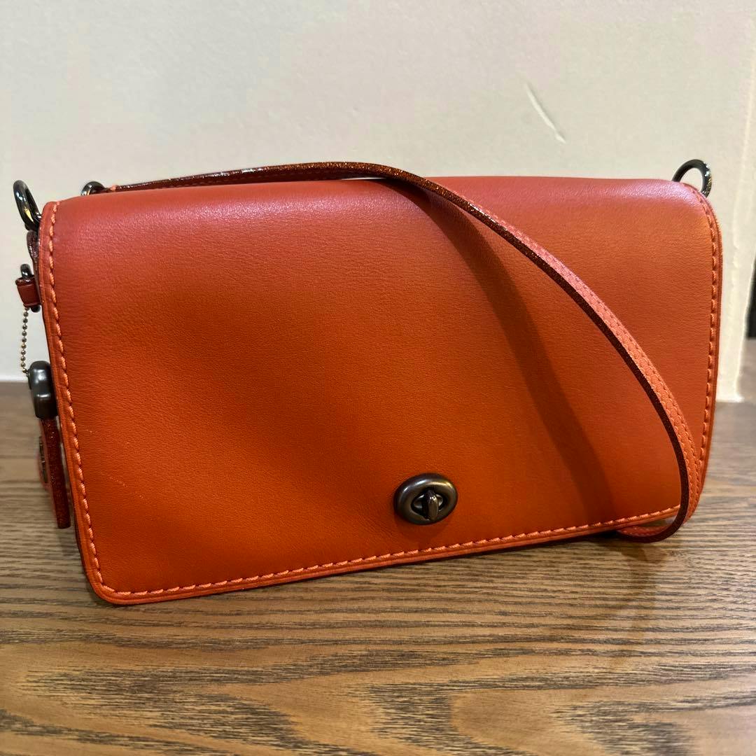 Front view of the red-orange Coach Dinky cow leather bag with strap and turnlock placed on a wooden surface.