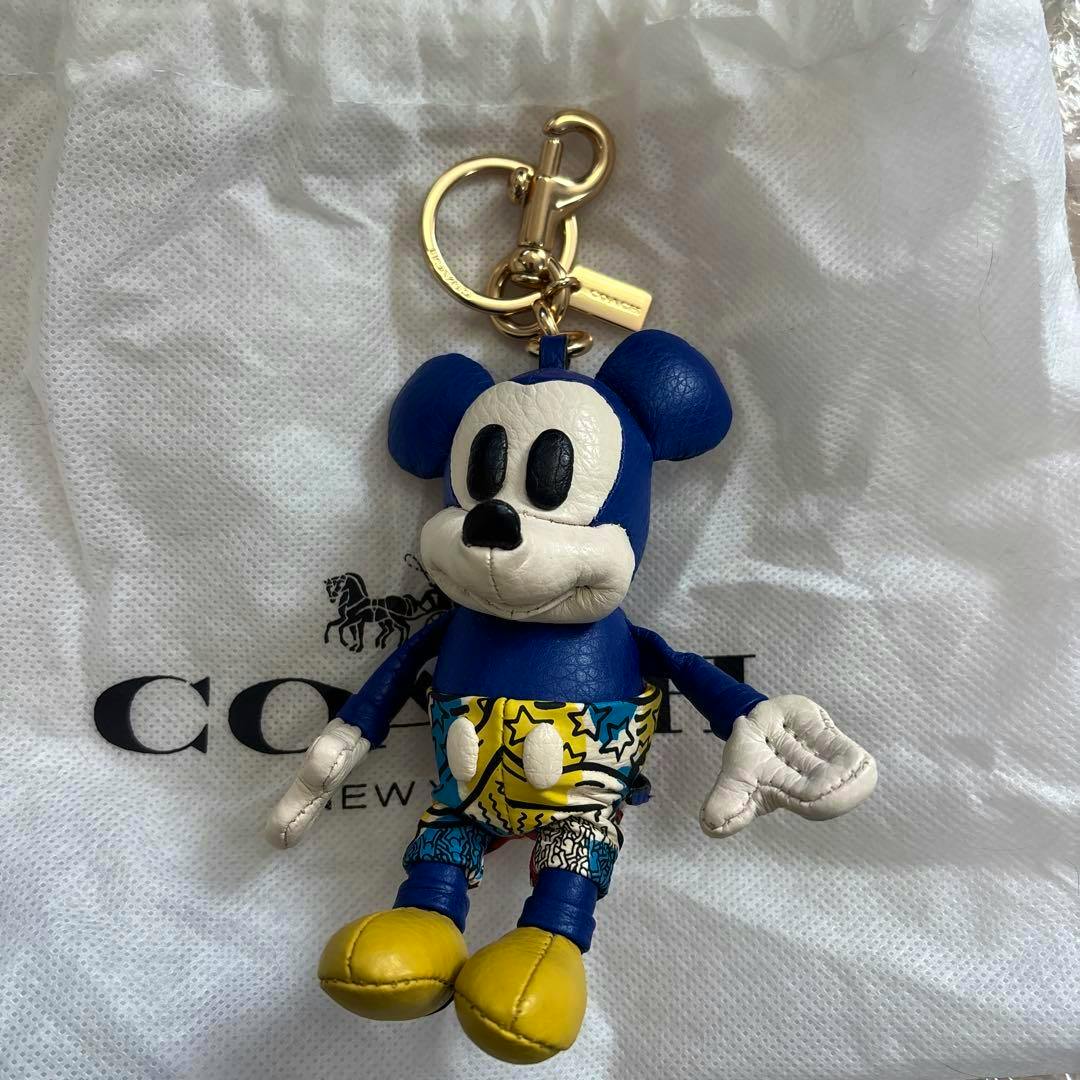 Front view of a Coach Disney bag charm shaped like Mickey Mouse, blue and white with patterned shorts and yellow shoes, photographed on a Coach dust bag.