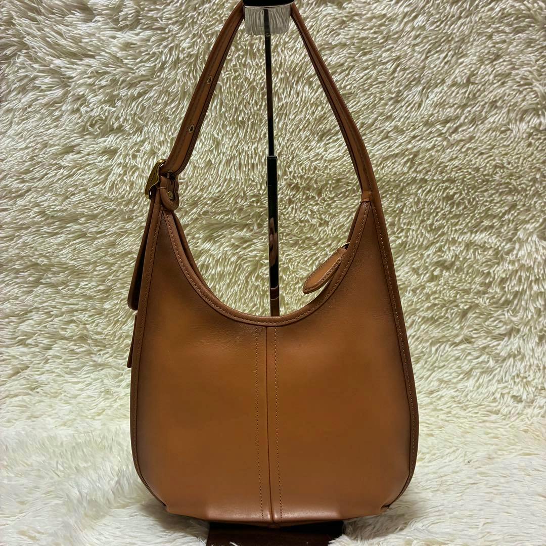 Brown Coach leather half-moon shoulder bag shown front view on a stand against a textured fabric background.