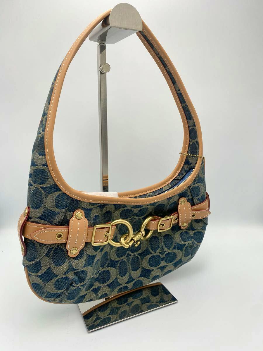 Front view of a Coach canvas shoulder bag with signature C pattern, tan leather trim, and brass belted hardware on a display stand.