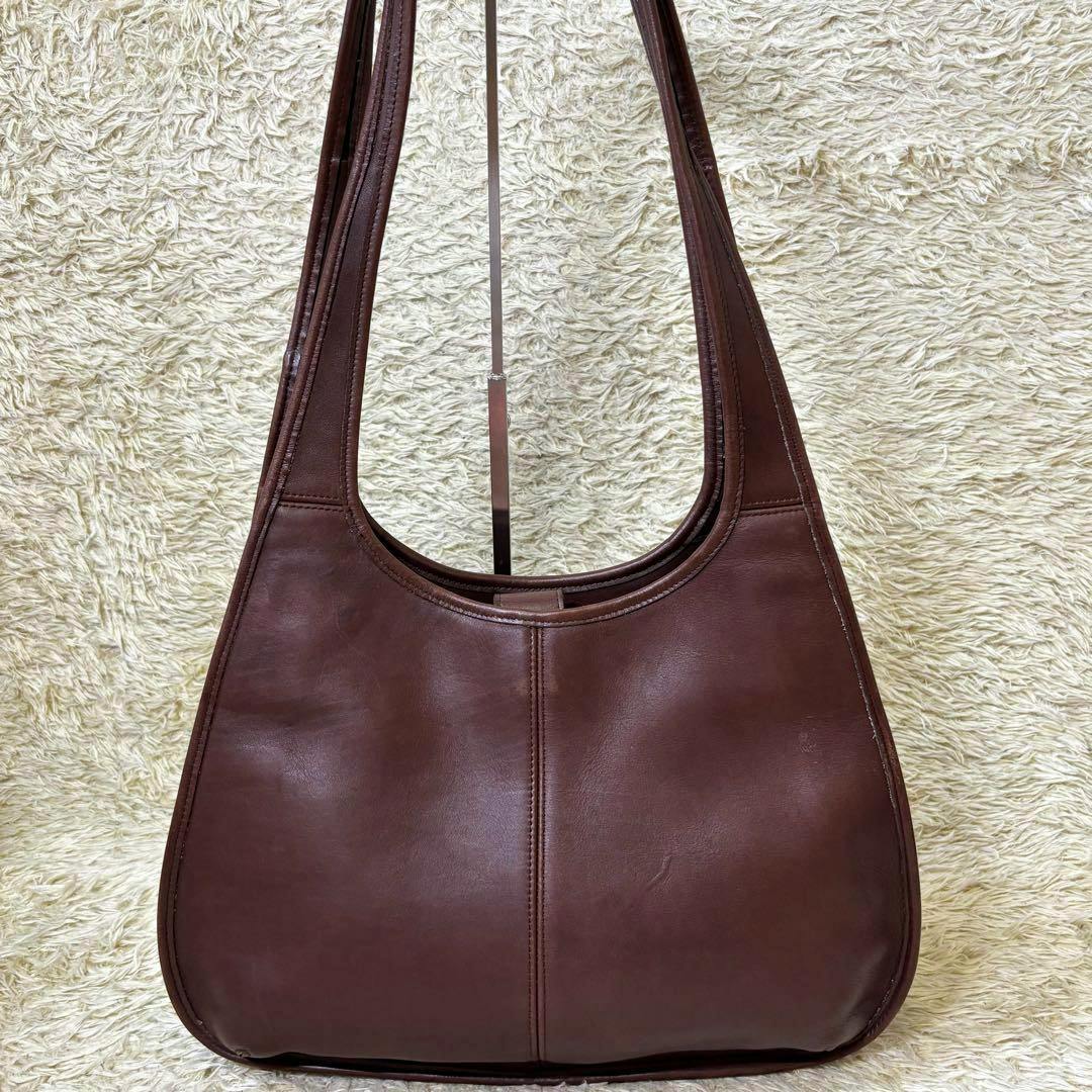 Front view of a mahogany Coach leather Ergo shopper bag hanging on a stand against a cream shag background.