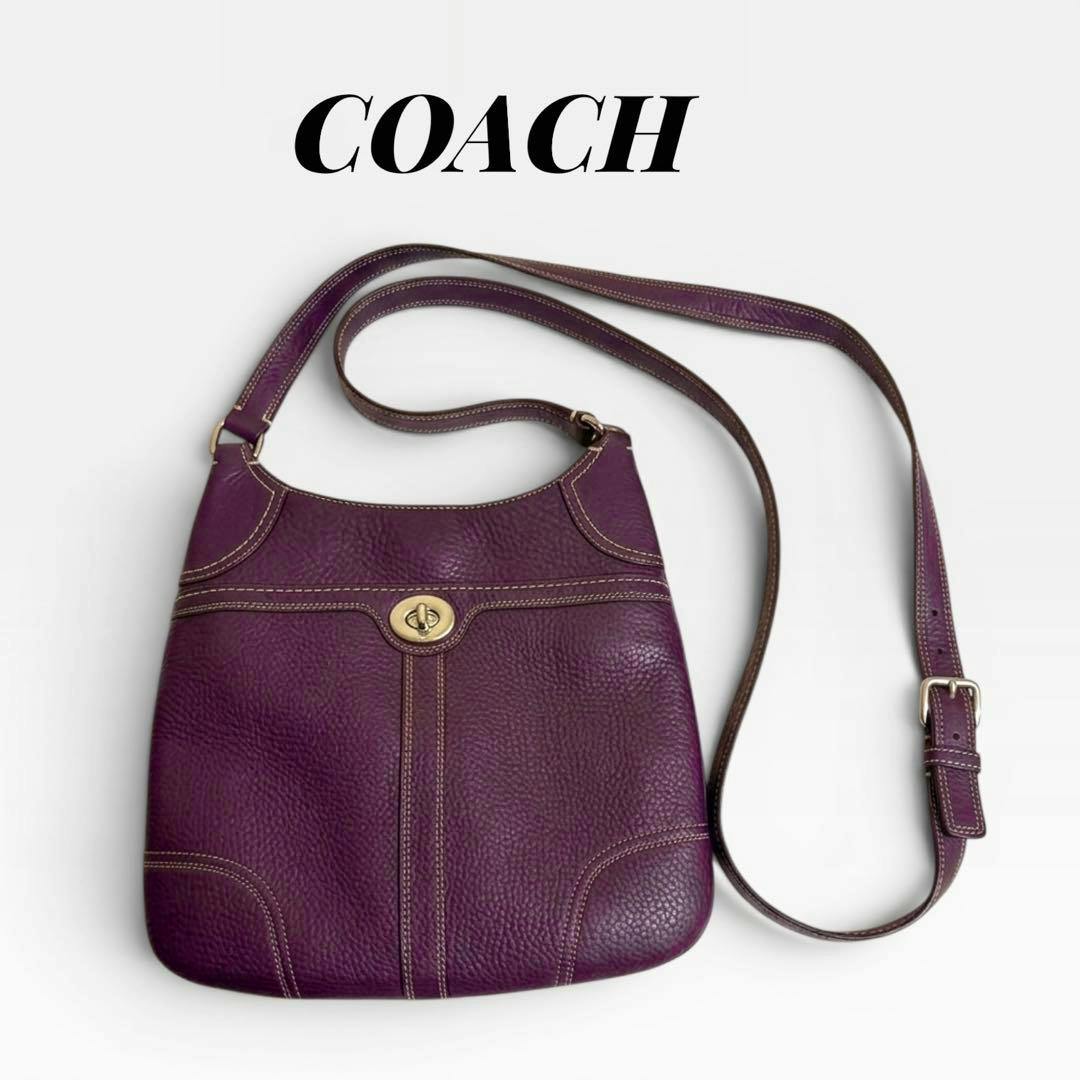 A purple Coach shoulder bag with a front turn-lock closure and a long adjustable strap on a white background.