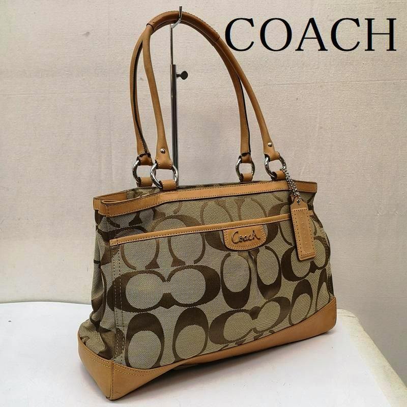 A Coach signature canvas tote bag with tan leather trim and shoulder straps displayed on a stand.