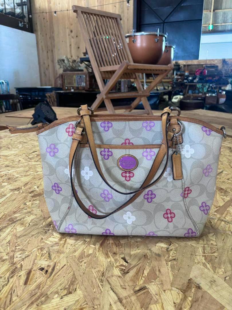 Front view of a COACH signature canvas floral-pattern tote bag with tan leather straps on a wooden surface.