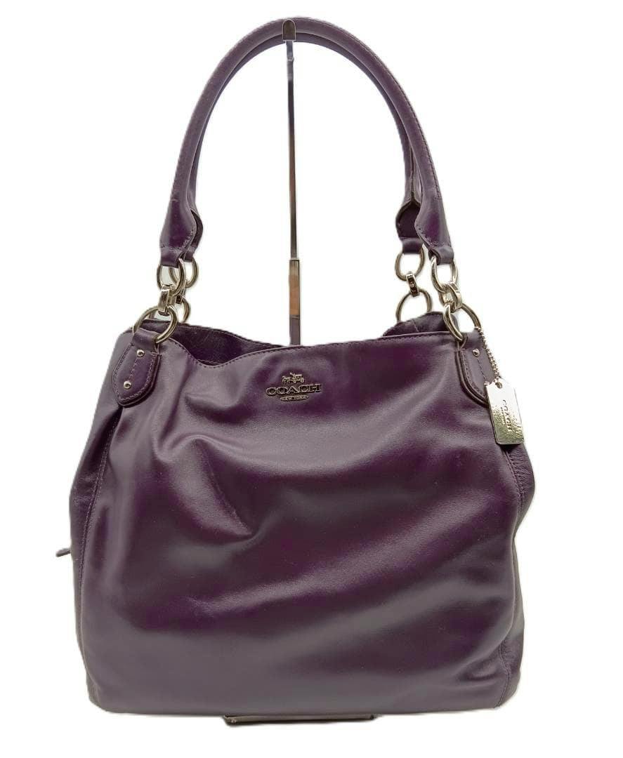Front view of the Coach leather shoulder bag showing top handles, metal Coach logo, and a hanging metal tag.