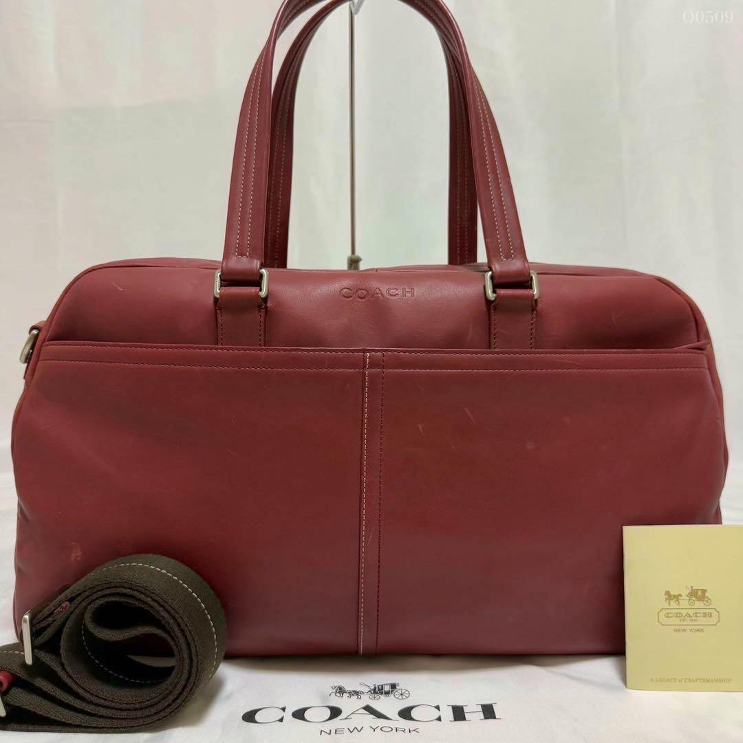 Front view of a red Coach leather 2-way Boston bag with top handles, a rolled shoulder strap beside it, and a Coach dust bag partially visible.