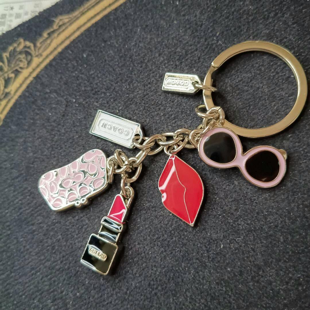 Coach keychain with pink-and-black sunglasses, red lips, red lipstick, pink patterned purse charm, and rectangular COACH tag on a dark fabric background.