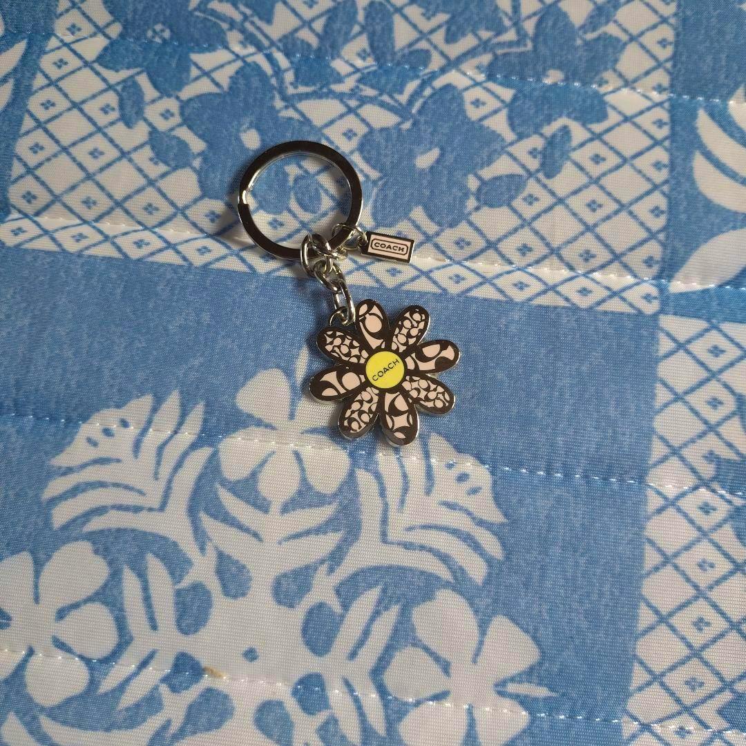 Front view of a COACH flower charm keychain with patterned petals and a yellow center on blue patterned fabric.