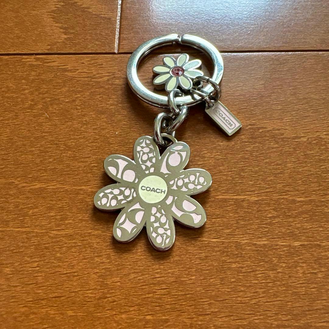 Front view of a COACH flower-motif metal keychain showing the daisy charm, small tag, and key ring on a wooden surface.