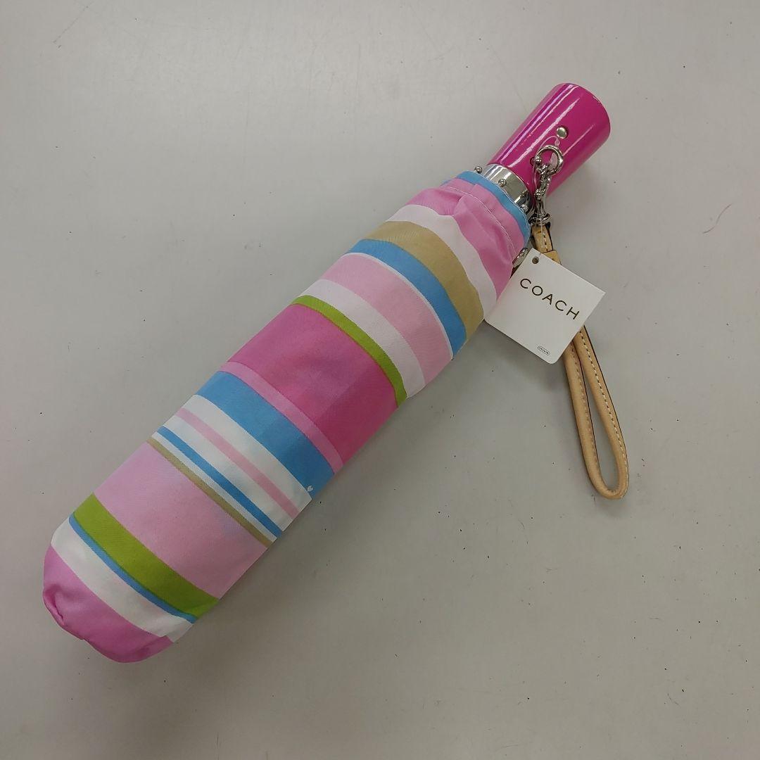 Folded striped pink COACH folding umbrella with wrist strap and attached tag on a light background.