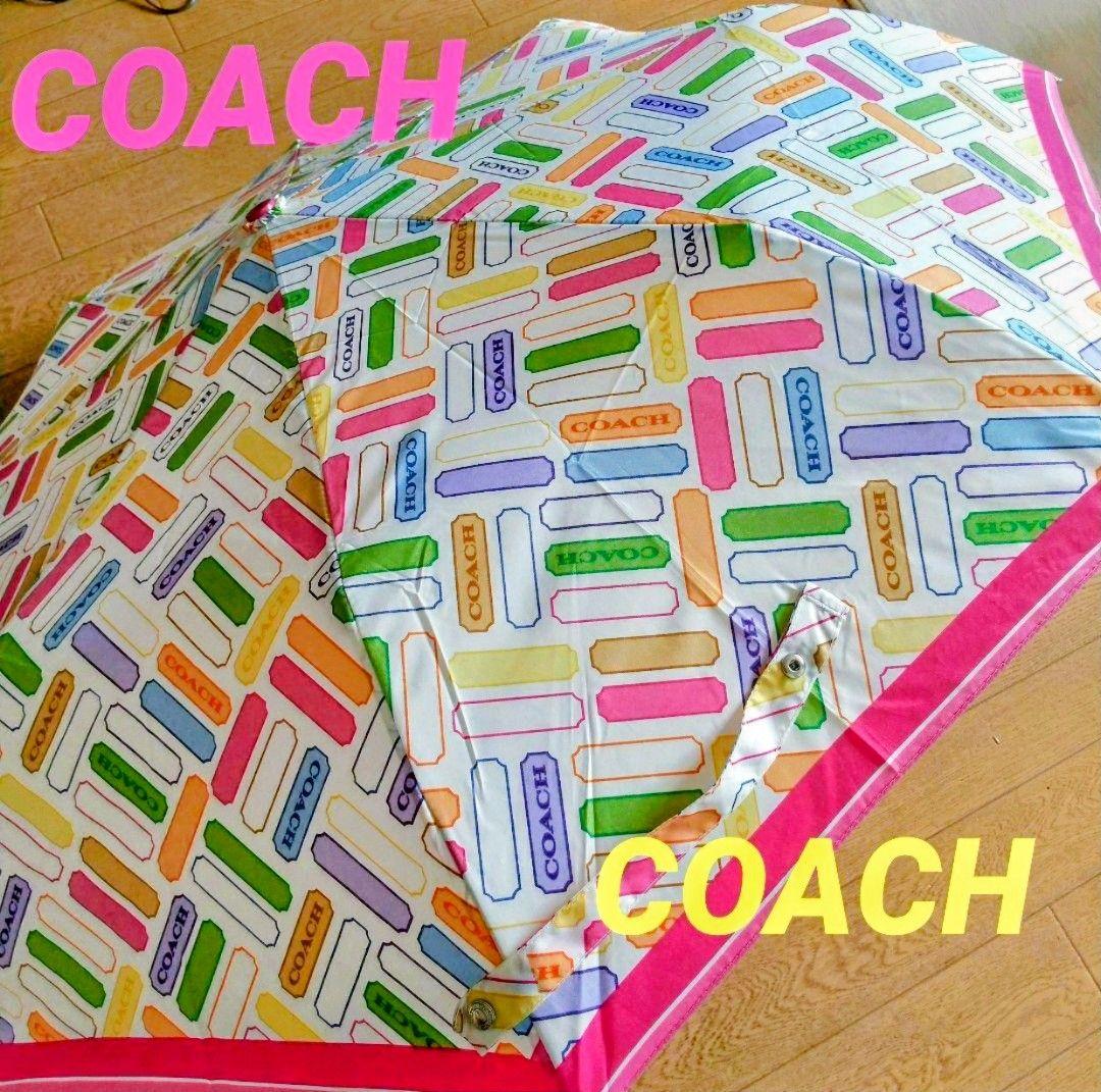 Open COACH folding umbrella with multicolored rectangular logo pattern and pink trim on a wooden floor.