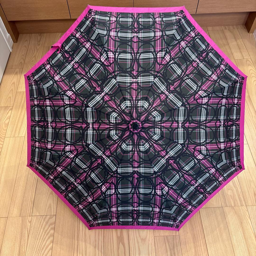 Open COACH folding umbrella with a black and pink checkered pattern seen from above on a wooden floor.