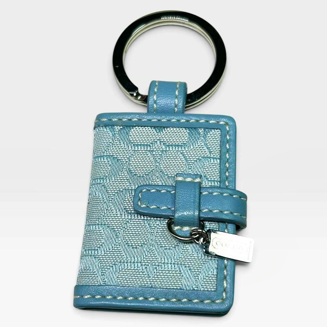 Blue book-shaped COACH key charm with stitched edges, a stitched strap closure, a small rectangular silver COACH tag, and a metal key ring on a white background.