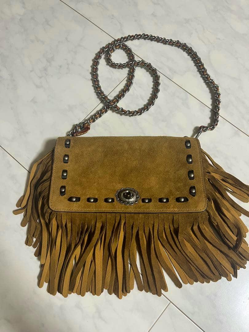 Brown suede Coach fringe chain shoulder bag laid flat showing the front flap with metal turnlock, stud detailing, long fringe, and chain strap.