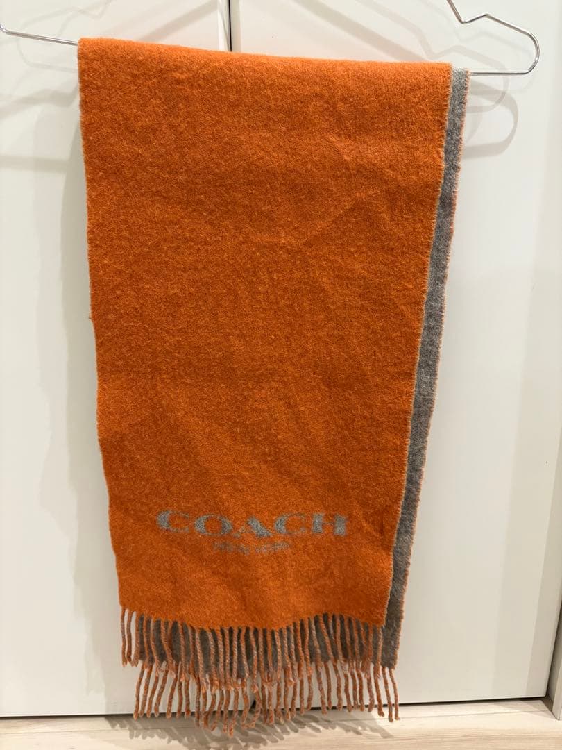COACH fringed wool muffler in orange and gray hanging on a hanger, showing the orange side with logo and fringed ends.