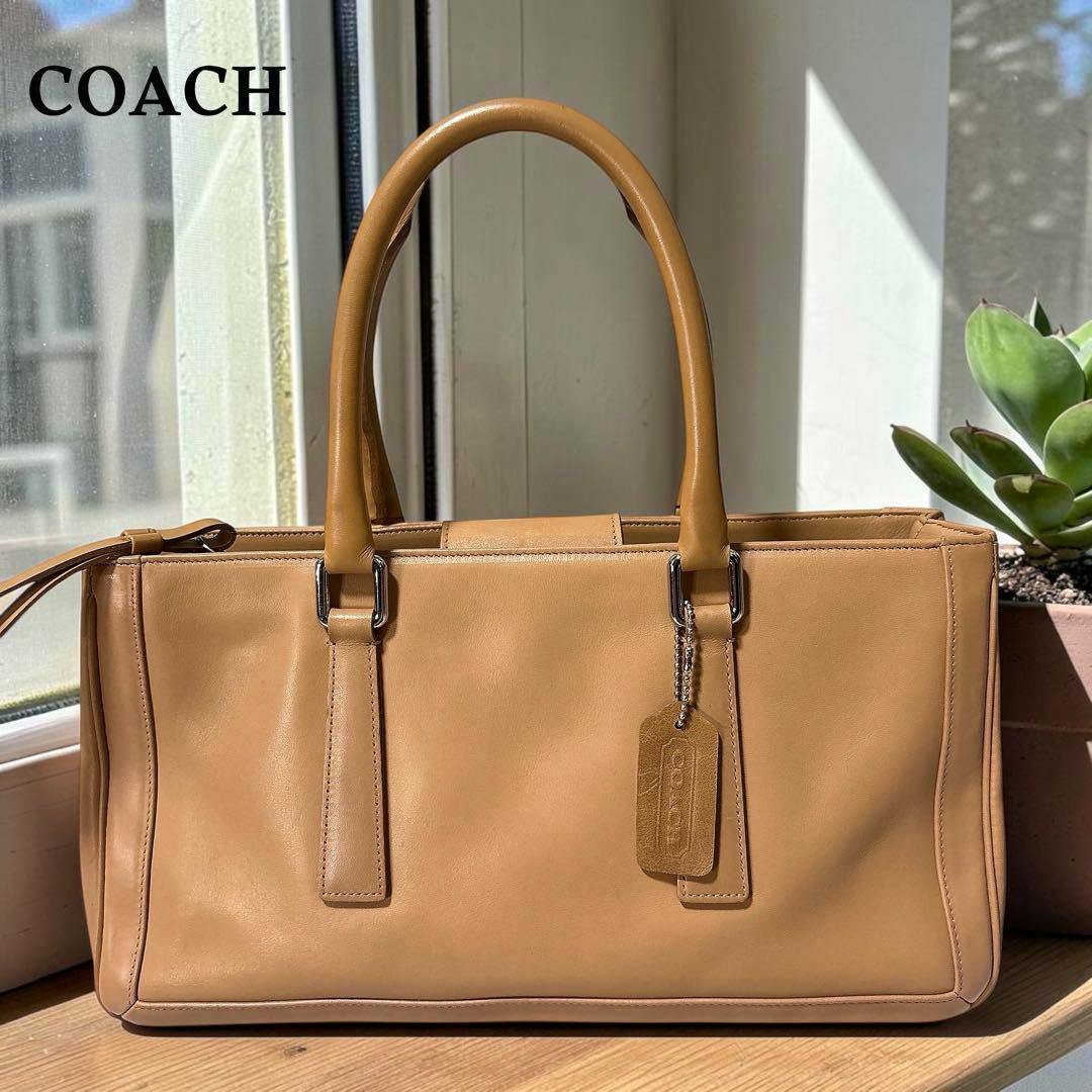Beige COACH leather handbag with dual top handles and a leather tag, photographed on a windowsill.