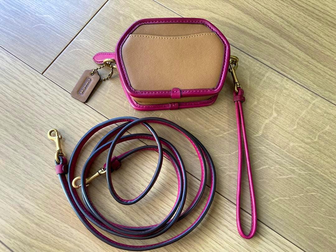 Tan and pink Coach coin case with a long detachable shoulder strap and wrist strap laid on a wooden floor.