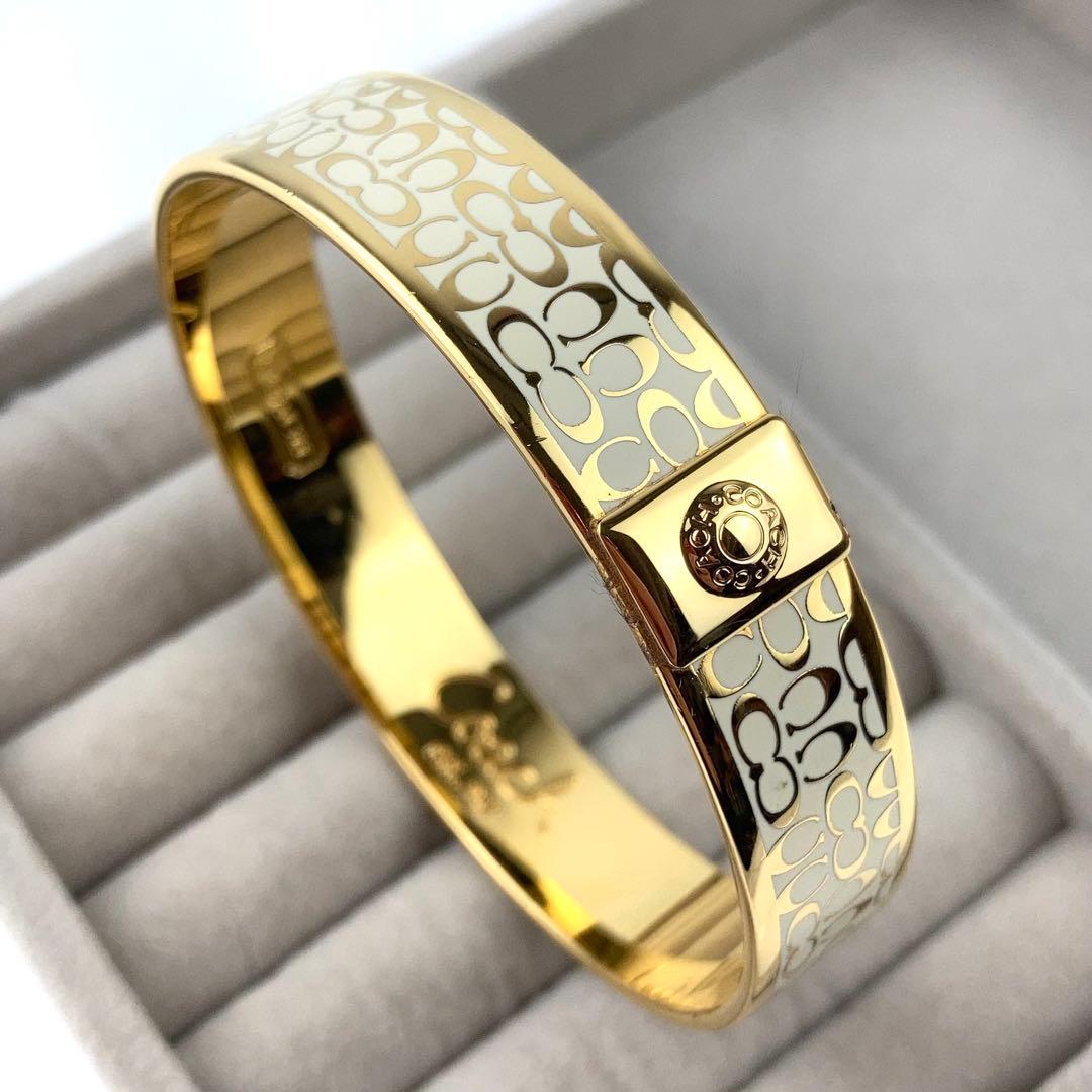 Gold Coach bangle bracelet with white signature 'C' pattern and clasp shown on a padded jewelry tray.