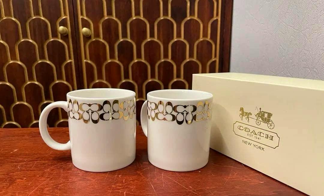 Two white Coach ceramic mugs with a gold pattern pictured on a table next to a Coach box.