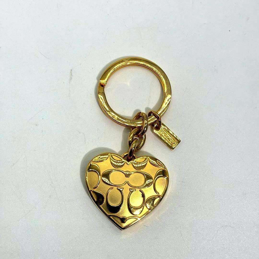 Gold heart-shaped bag charm keychain with embossed signature pattern and a small rectangular tag on a white background.
