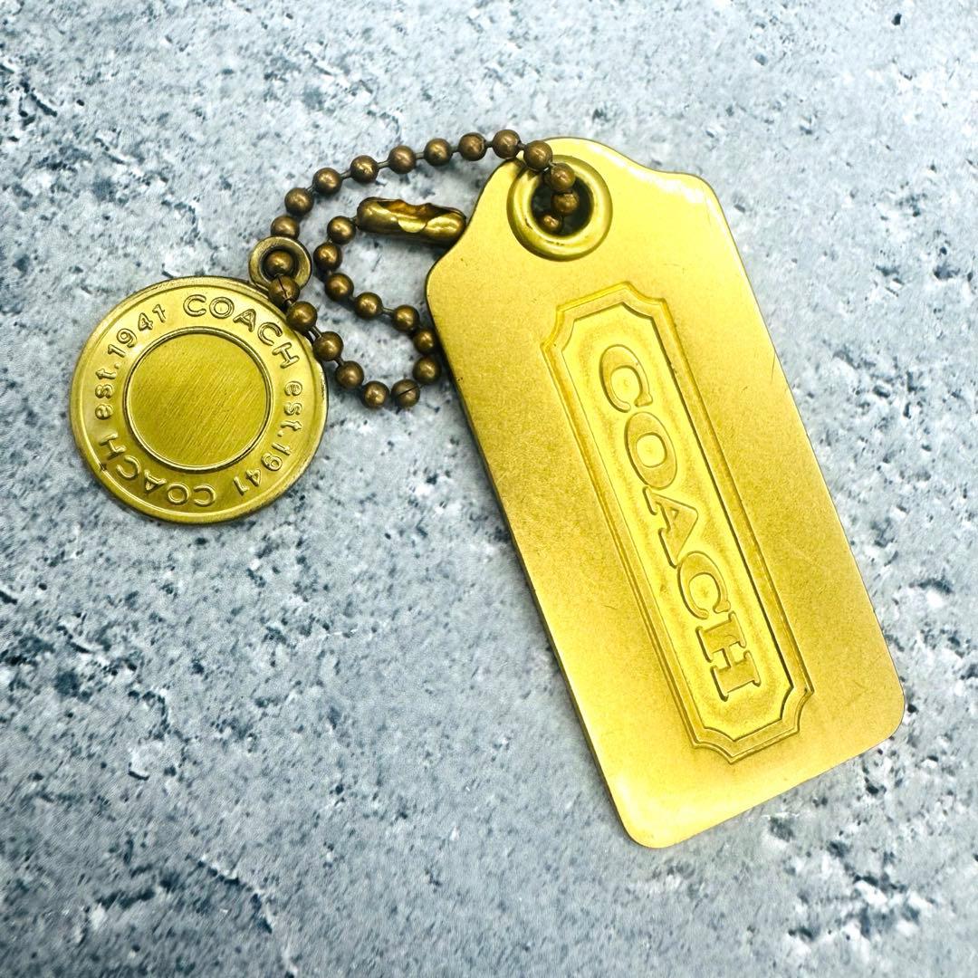 Gold COACH rectangular tag keychain with a round 1941-logo medallion displayed on a textured gray surface.