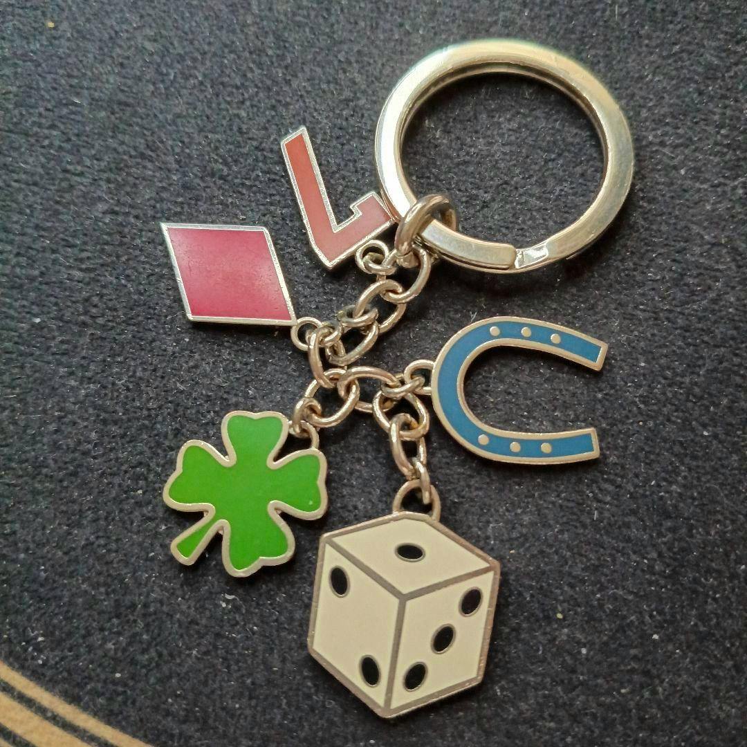 A zinc-alloy keychain on a dark surface showing multiple luck-themed charms — dice, four-leaf clover, horseshoe, and a red diamond-shaped charm.