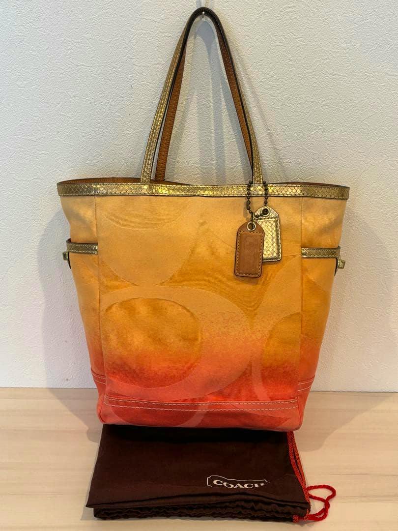 Front view of an orange gradient Coach canvas tote bag with metallic trim, leather tags, and a Coach dust bag underneath.