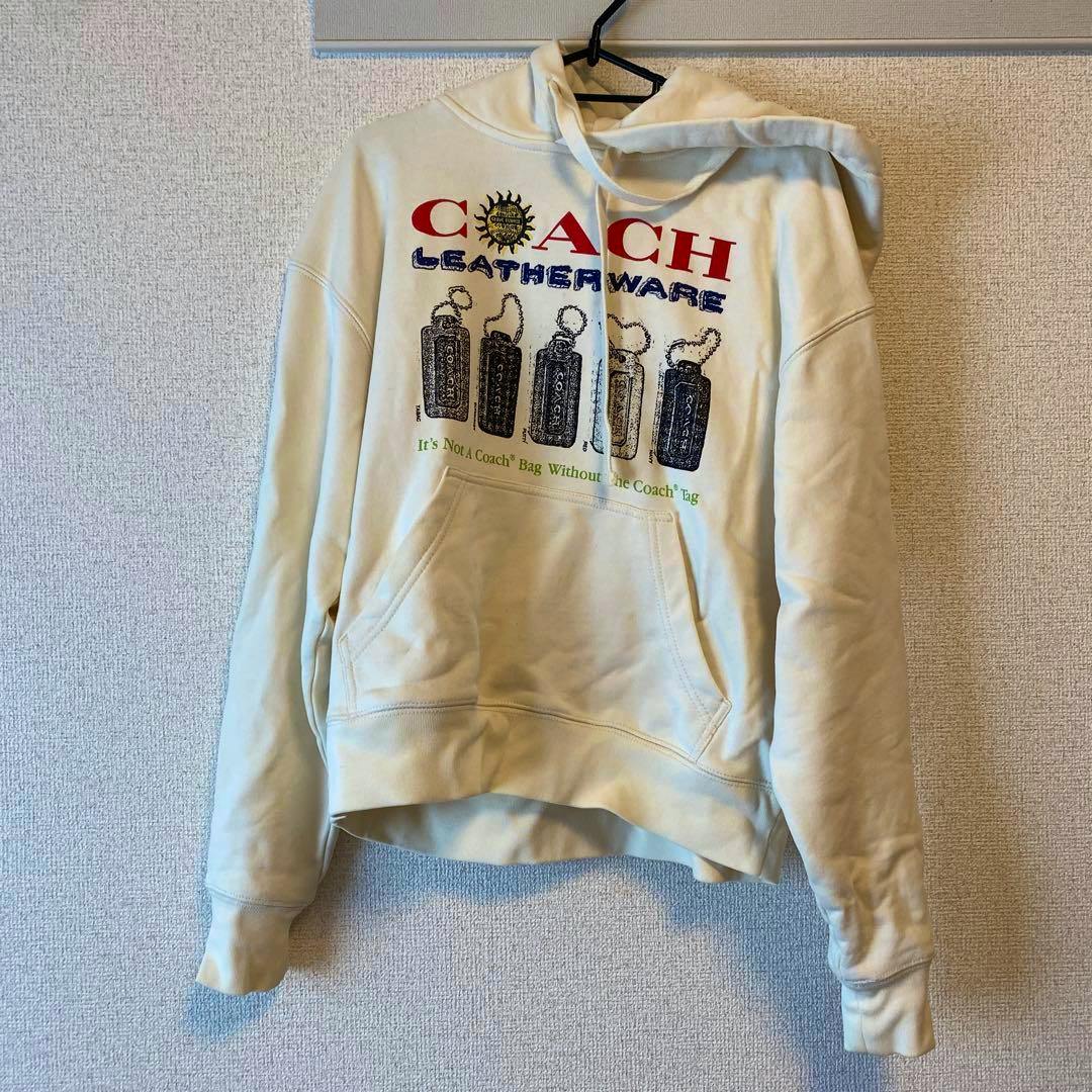 Cream COACH graphic hoodie hanging on a wall, front view showing printed logo and kangaroo pocket.