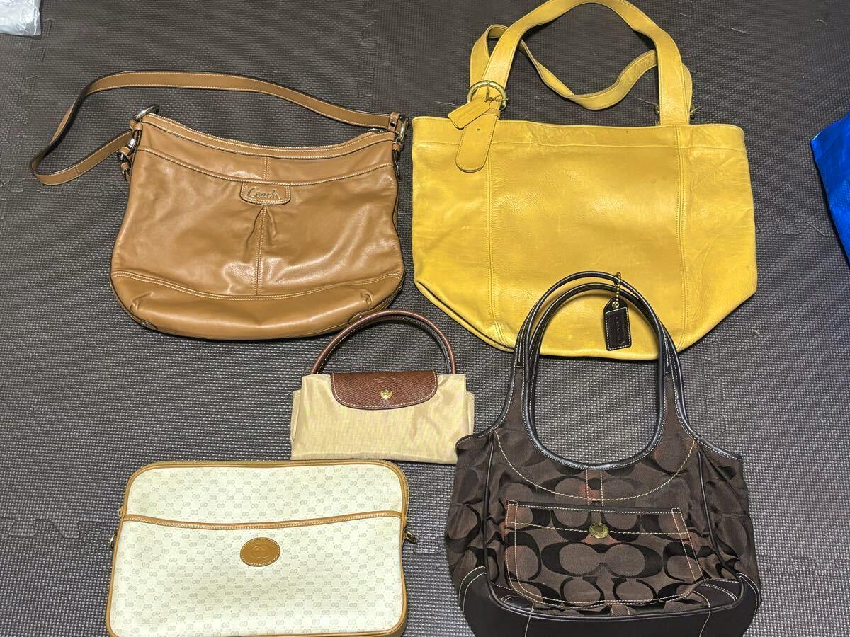 Five bags arranged on a floor mat: a brown leather Coach shoulder bag, a yellow leather tote, a small beige handbag, a white patterned pouch, and a brown Coach monogram tote.