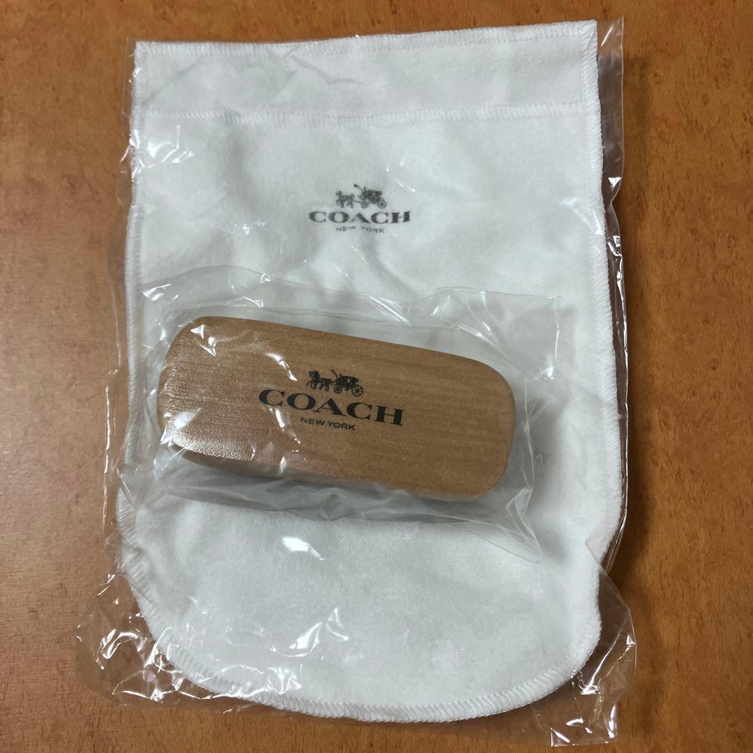 A wooden COACH hair brush wrapped in plastic resting on a white COACH pouch, both in clear plastic packaging on a wooden surface.