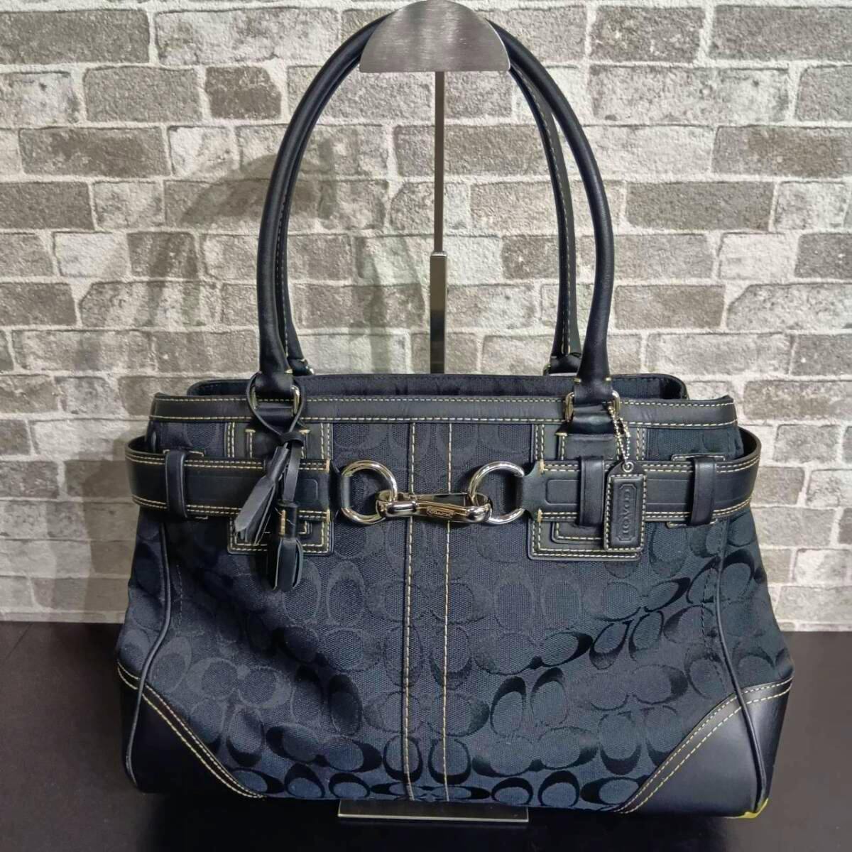 Front view of a Coach signature canvas handbag with leather trim, double handles, and metal bit hardware shown on a display stand.