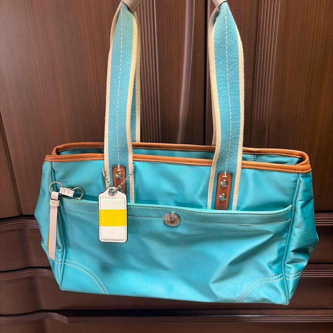 Light-blue COACH Hamptons satin satchel tote bag with striped canvas shoulder handles, brown leather trim, front slip pocket, and a hanging white-and-yellow tag.