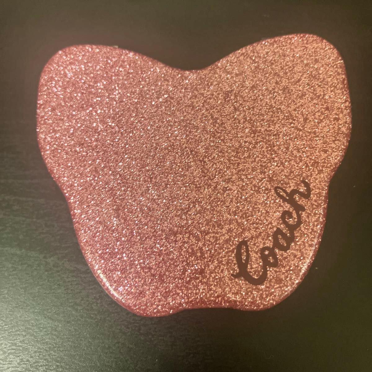 Pink glitter Coach compact hand mirror with the Coach logo on the front, shown closed on a dark surface.