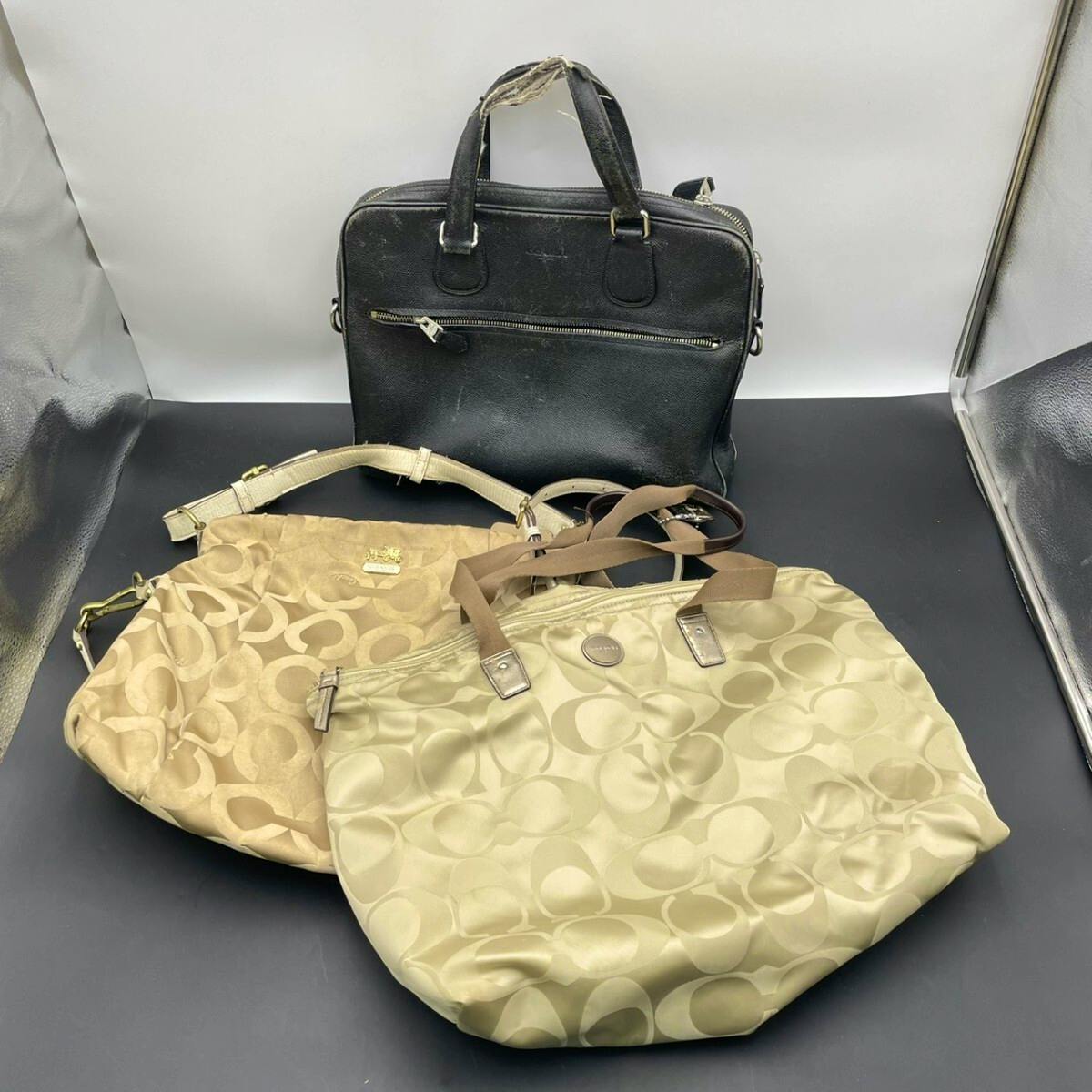 Three bags: a black handled Boston-style bag and two beige Coach monogram tote bags displayed on a black surface.