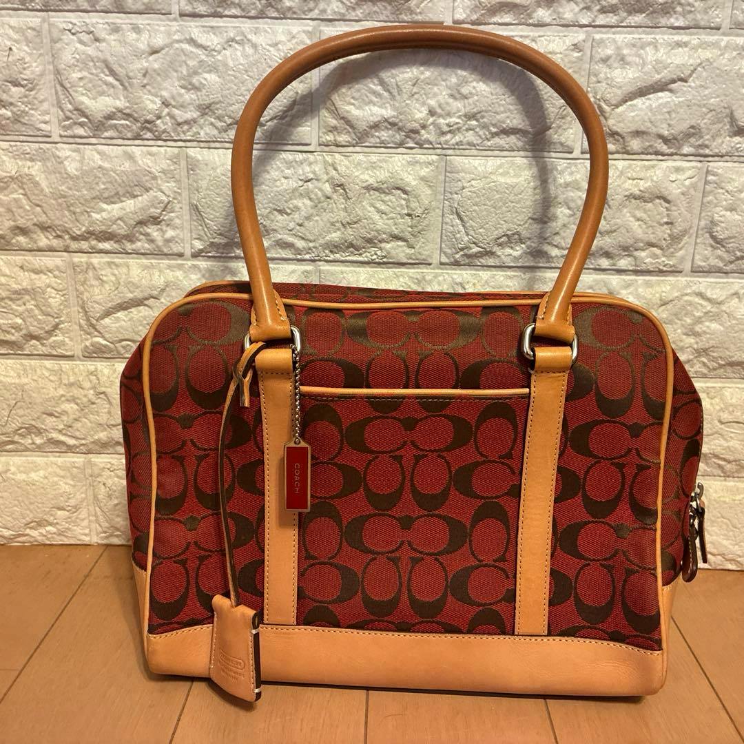 Front view of a brown/beige COACH canvas handbag with leather handles and signature C pattern, placed on a wooden floor.