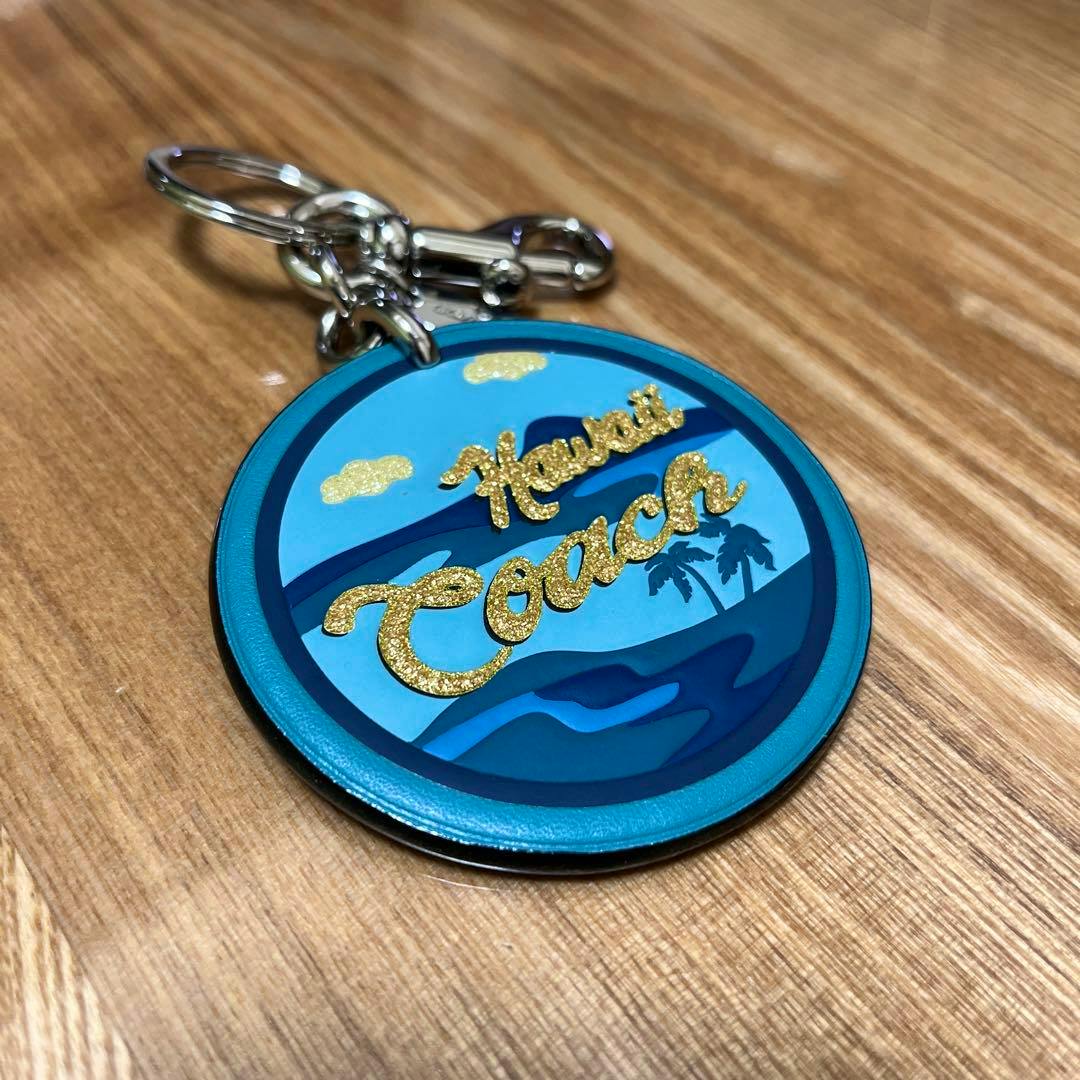 Round blue COACH Hawaii bag charm with gold glitter "Hawaii Coach" script lying on a wooden surface.