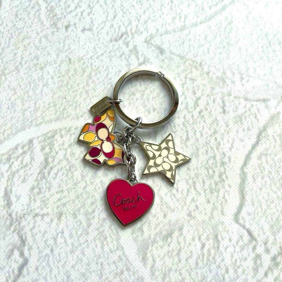 A Coach keyring with a pink heart charm, a multicolored butterfly charm, and a star charm on a white textured background.