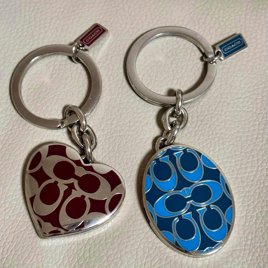 Two COACH keychains with enamel signature-C patterns, one heart-shaped in maroon and one oval in blue, shown on a light textured surface.