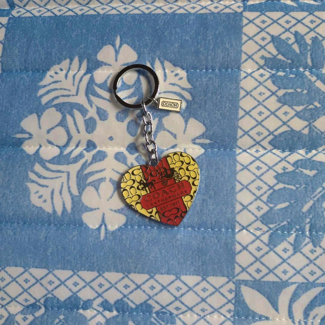 COACH heart-shaped metal keychain with a red center and yellow patterned front attached to a metal ring, shown on blue patterned fabric.