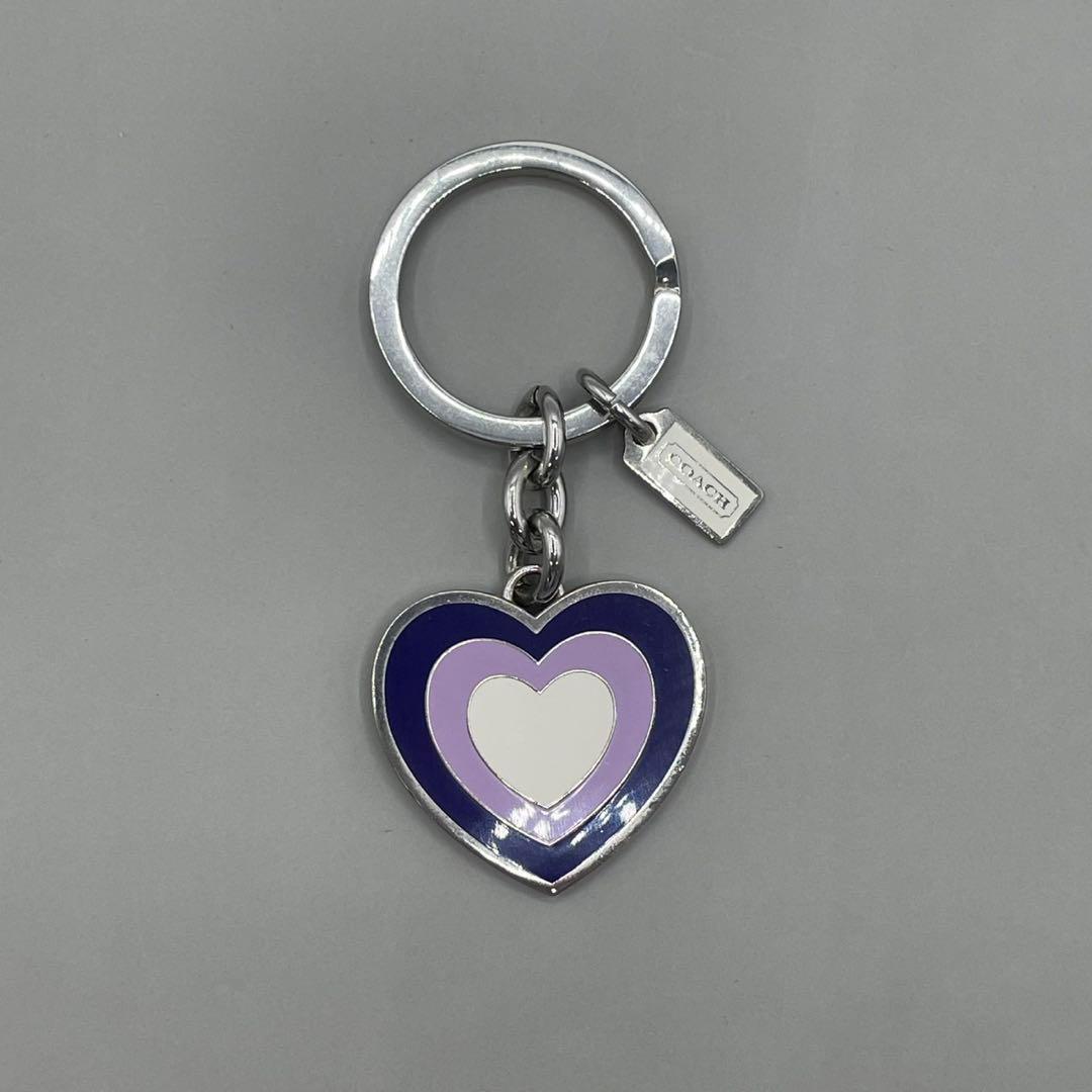 Silver keyring bag charm with a purple, lavender, and white concentric heart enamel pendant and a small Coach tag on a gray background.