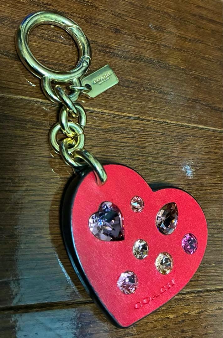 Red COACH heart leather keychain charm with gold-tone ring and multicolored crystal studs on a wooden surface.