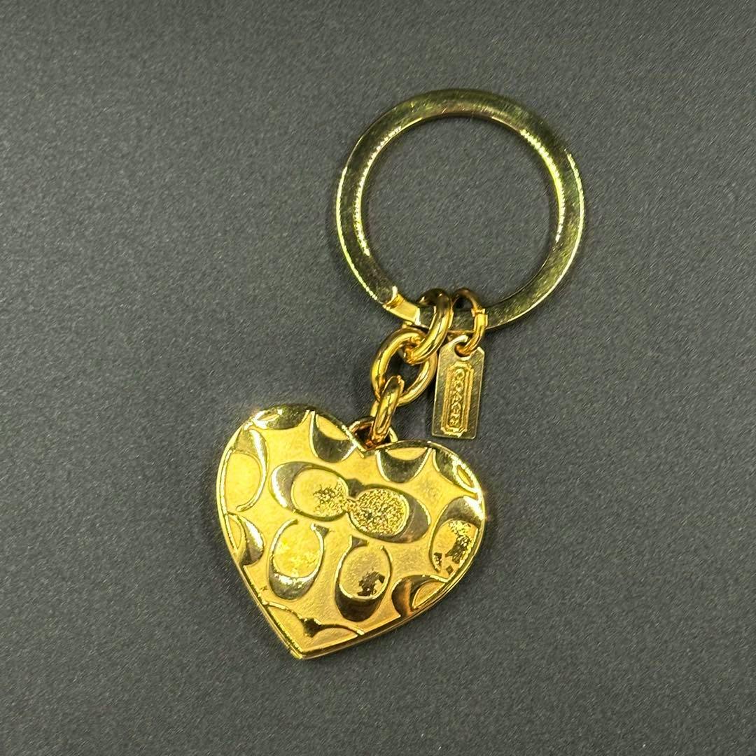 Gold heart-shaped locket key charm with Coach signature pattern, small logo tag, and round key ring on a gray background.