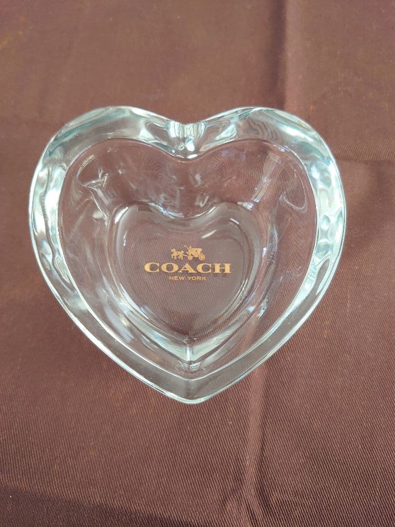 Top-down photo of the COACH heart-shaped clear glass ashtray/small dish showing the gold COACH logo on a brown background.