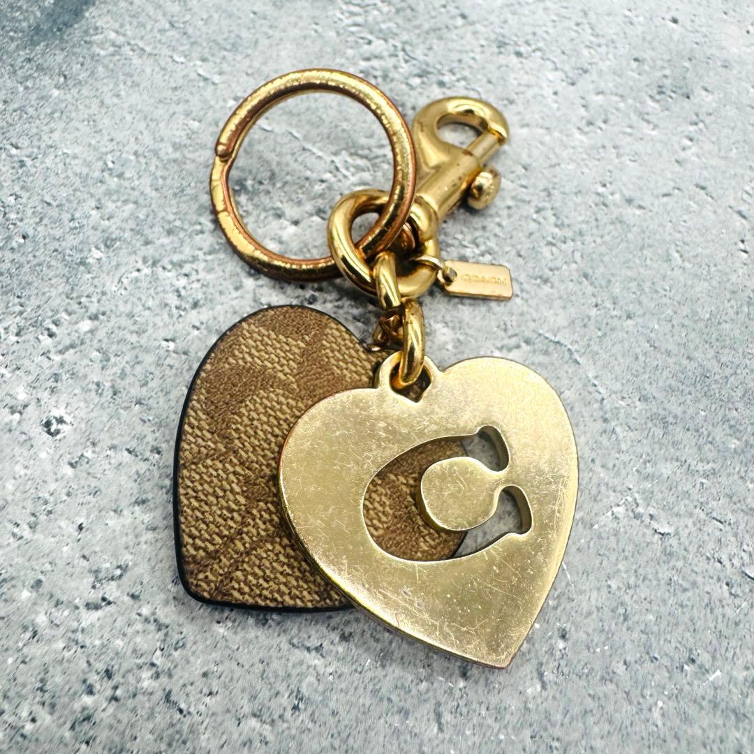 COACH heart-shaped keychain showing a gold metal heart with a cut-out 'C' logo and a brown patterned heart on a gray background.