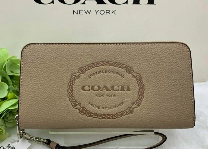Long zip-around COACH leather wallet with embossed Coach logo on the front and attached wrist strap, shown against a COACH backdrop.